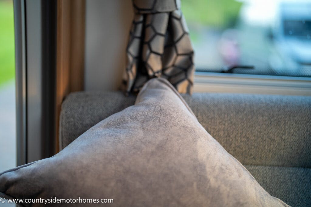 A close-up photograph of a gray pillow placed against a gray upholstered seat in the 2021 Bailey Autograph 79-4i motorhome. In the background, there is a window with a curtain featuring a geometric pattern. Part of the outdoor scenery is slightly visible through the window.