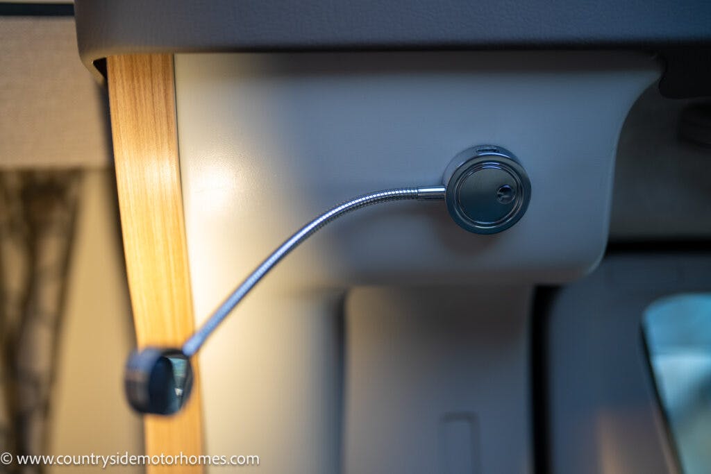 A close-up of a flexible gooseneck reading light mounted on the interior wall of what appears to be a 2021 Bailey Autograph 79-4i motorhome. The light, with its small circular head, is positioned next to a wooden panel and is switched off.