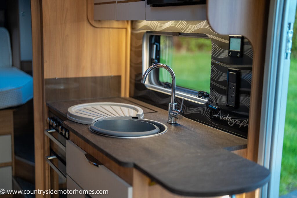 The image shows the kitchen area of a 2021 Bailey Autograph 79-4i motorhome with a sleek countertop, oval sink, modern faucet, and built-in stove. The backsplash has a wavy design and a small window. There's a control panel on the wall, and storage cabinets are visible above and below.