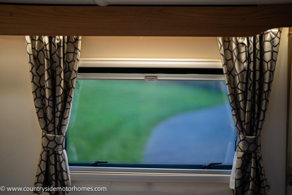 A window in the 2021 Bailey Autograph 79-4i motorhome with patterned curtains pulled to the sides. The view outside shows a blurry green field and a pathway. The wooden frame above the window matches the interior decor, enhancing the cozy ambiance.