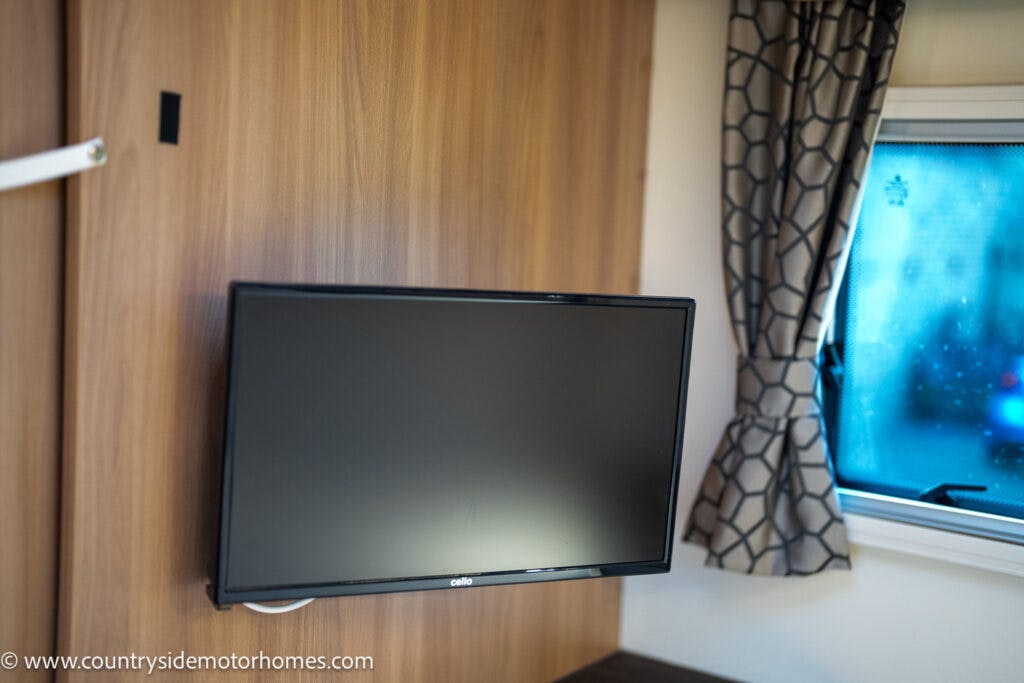 A flat-screen television is mounted on a wooden wall next to a window with patterned curtains. The television screen is turned off, and the URL www.countrysidemotorhomes.com is visible at the bottom left of the image, showcasing their 2021 Bailey Autograph 79-4i model.