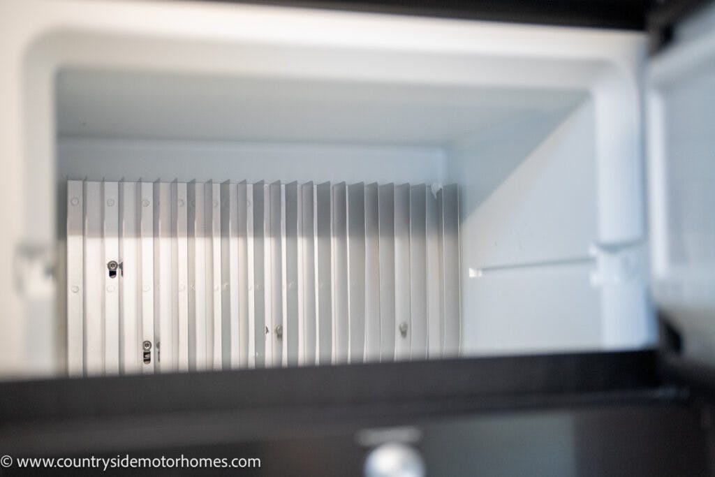 The image shows the interior of an empty refrigerator or freezer compartment, likely from the 2021 Bailey Autograph 79-4i. The walls are light grey with ribbed detailing on the back. There are no shelves or contents inside, and the door is partially open. A web address is visible in the bottom-left corner.
