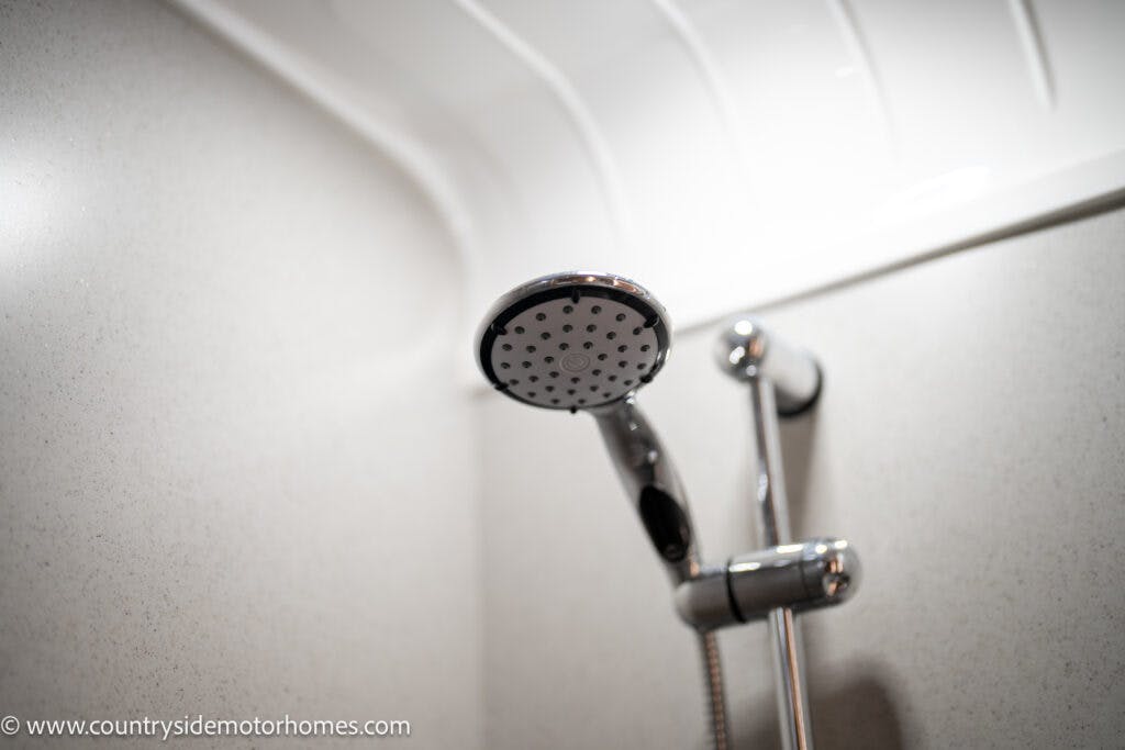 Close-up of a shower head in the 2021 Bailey Autograph 79-4i bathroom setting. The shower head, mounted on a wall with a shiny chrome finish, is adjustable in height. The background showcases white, textured wall panels and a slightly curved ceiling.