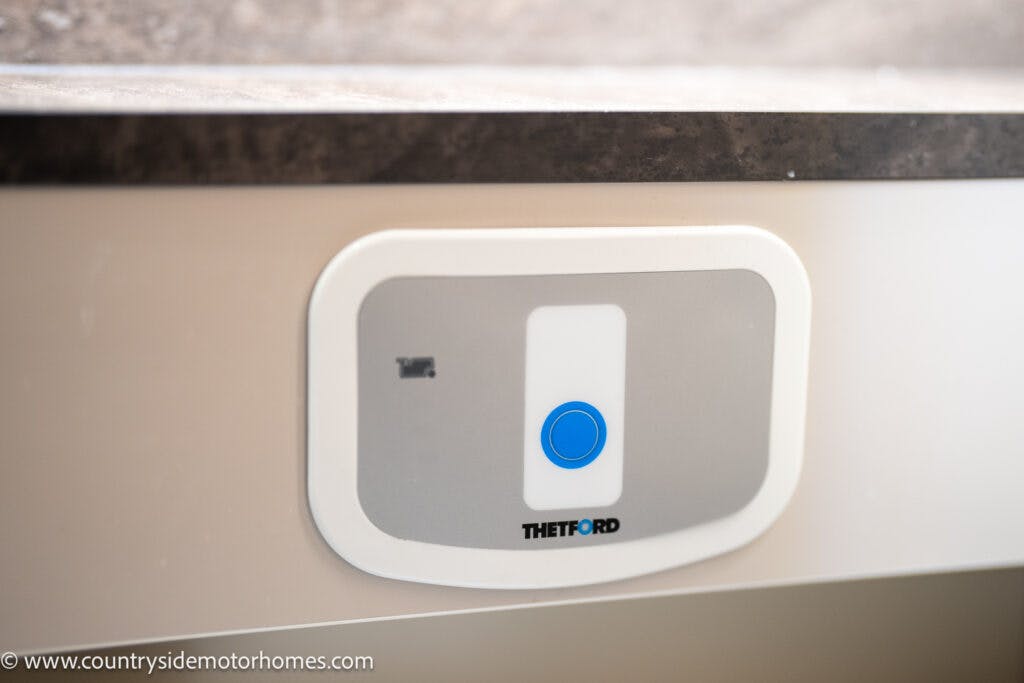 A close-up view of a control panel with a blue button and an indicator light, labeled "THETFORD" inside the 2021 Bailey Autograph 79-4i. The panel is housed in a light-colored plastic casing and is installed on a flat surface. Image © countryside motorhomes.