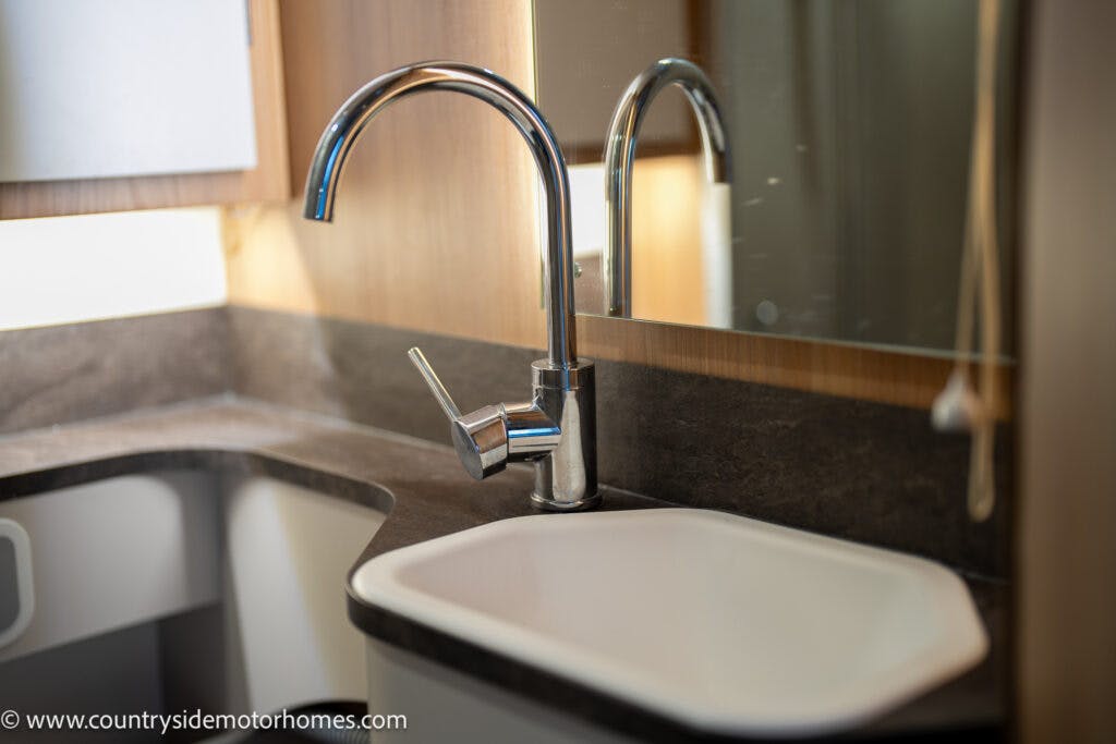 A close-up view of a modern kitchen sink with a sleek, curved faucet, featured in the 2021 Bailey Autograph 79-4i. The sink is set in a countertop with a dark finish. There is a mirror and light wooden cabinetry in the background. A web address, www.countrysidemotorhomes.com, is visible in the lower left corner.