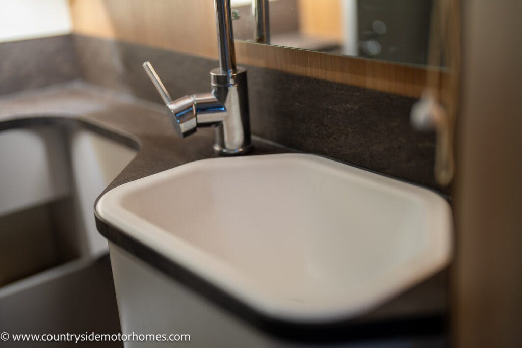 A close-up view of a white rectangular sink with a modern chrome faucet in the 2021 Bailey Autograph 79-4i. The sink is set into a dark countertop, with a mirrored backsplash above. The left side of the image shows part of an adjacent counter or cabinet.