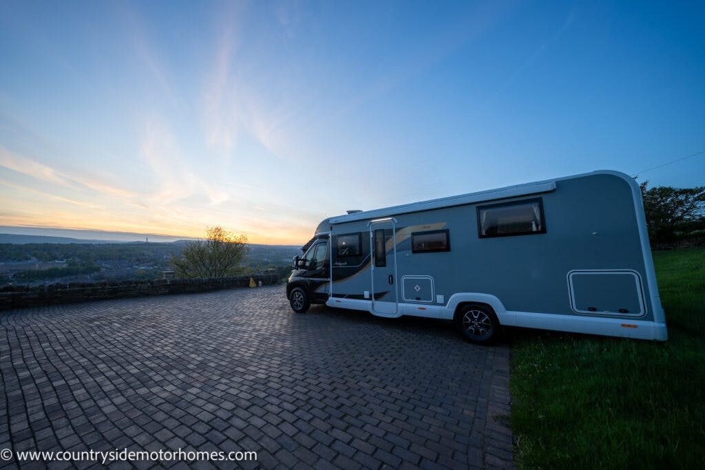A 2021 Bailey Autograph 79-4i RV is parked on a paved area overlooking a scenic landscape at dusk. The sky is clear with a subtle gradient from light blue to soft yellow. There is a grassy area to the right and a distant view of trees and hills.