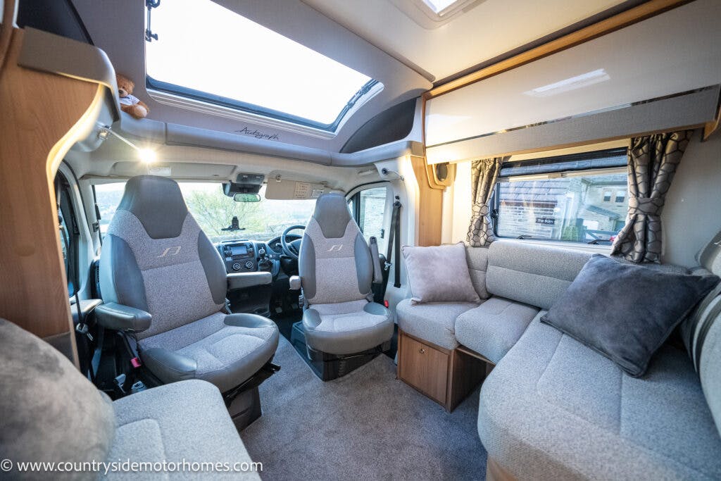 Interior view of the 2021 Bailey Autograph 79-4i motorhome featuring a seating area with cushioned benches and throw pillows, captain's chairs at the front, a skylight, and large windows with curtains. The space is designed for comfort and travel.