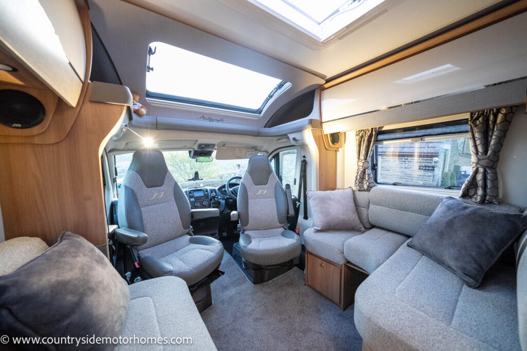 Interior view of the 2021 Bailey Autograph 79-4i campervan featuring a driver's seat and a passenger seat in the front, with a spacious seating area including a sofa and a small table. The space is well-lit with natural light from a sunroof. Upholstery is in grey fabric.