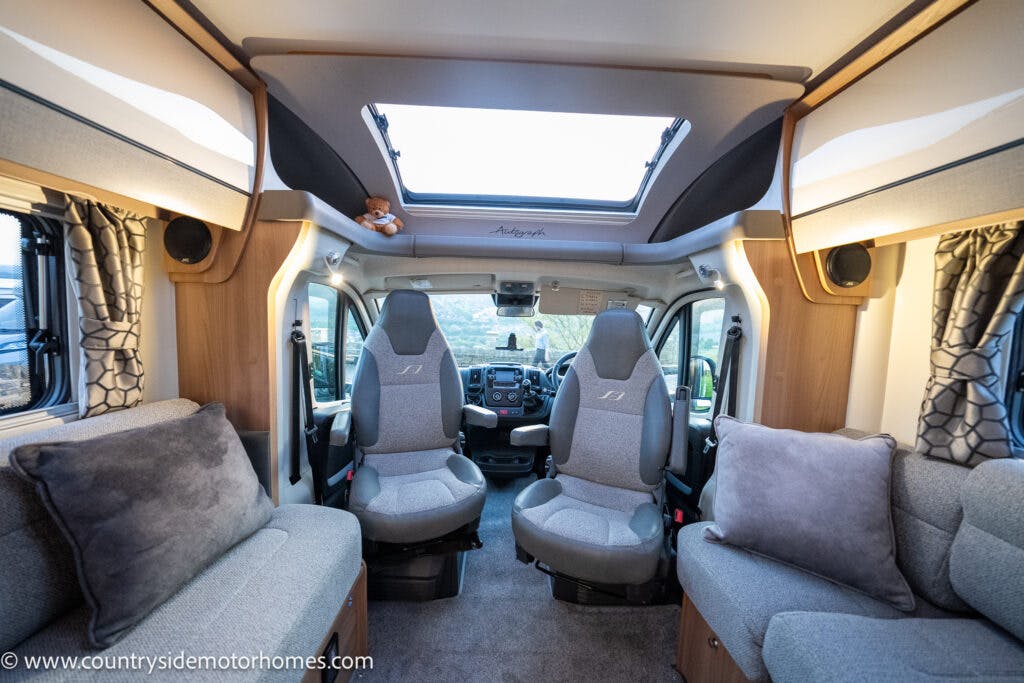 The interior of the 2021 Bailey Autograph 79-4i motorhome features two front seats facing the living area. Windows and curtains are visible on both sides. The furnishings include grey upholstery, a small teddy bear on a shelf, and pillows on the couch. The skylight allows natural light in.
