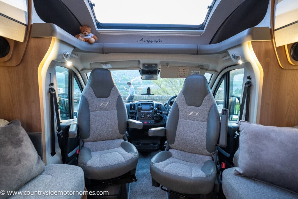 The interior of the Bailey Autograph 79-4I motorhome features two grey captain chairs at the front, a steering wheel, and a dashboard. A skylight is visible above, and there is a teddy bear placed on a shelf. Through the windows, outdoor greenery can be seen.

