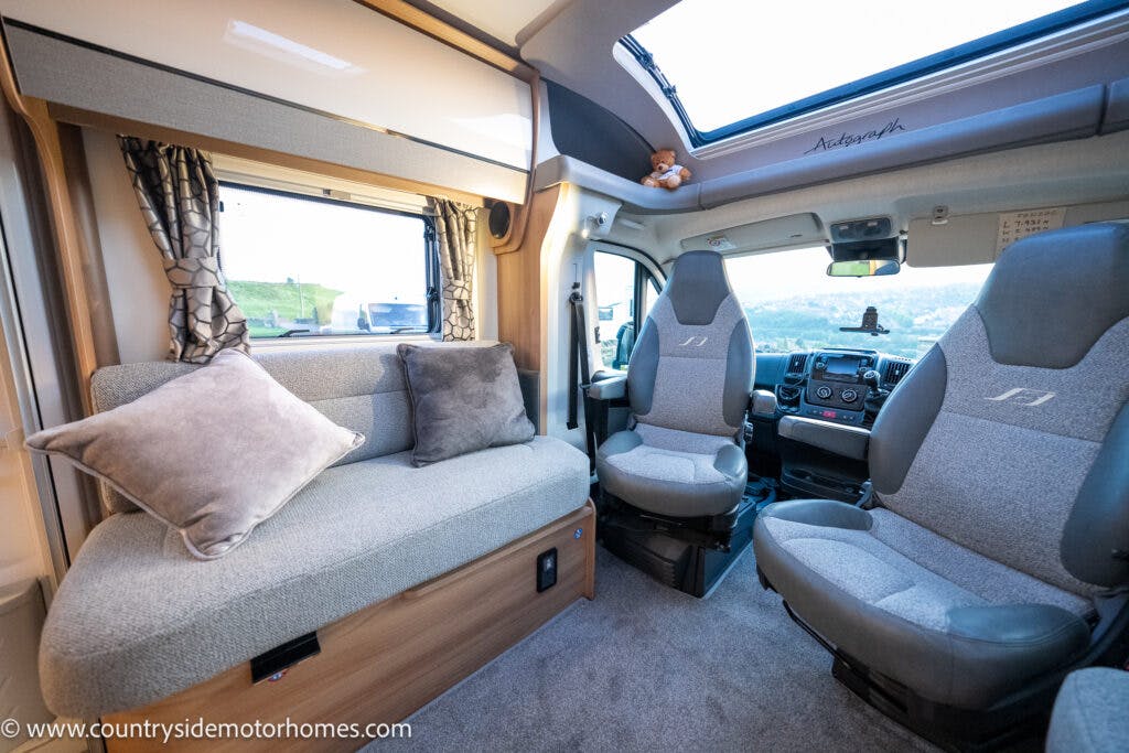 The interior of the 2021 Bailey Autograph 79-4i motorhome features a grey cushioned bench with two pillows on the left, two grey captain's chairs in the front, a large window with curtains, and a view of the exterior landscape.