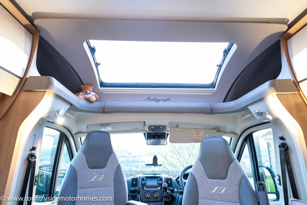 The interior view of the 2021 Bailey Autograph 79-4i motorhome showcases the driver's and passenger's seats, a panoramic sunroof with a teddy bear perched on the edge, and various controls on the dashboard. The steering wheel features the vehicle manufacturer's logo, and the windscreen offers a clear outside view.