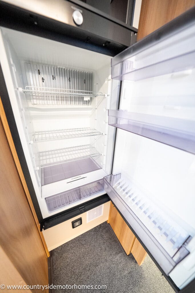 Open, empty refrigerator with several shelves and door compartments visible inside a 2021 Bailey Autograph 79-4i. The interior is clean and well-lit, nestled in a wooden kitchen unit. The floor is carpeted. A website URL is visible at the bottom left of the image.