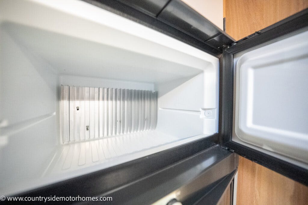 An open, empty mini fridge is shown with its door fully open. The interior is clean and features a ridged area on the back wall, likely for cooling purposes. To the right, the edge of a wooden cabinet from the 2021 Bailey Autograph 79-4i motorhome is visible.