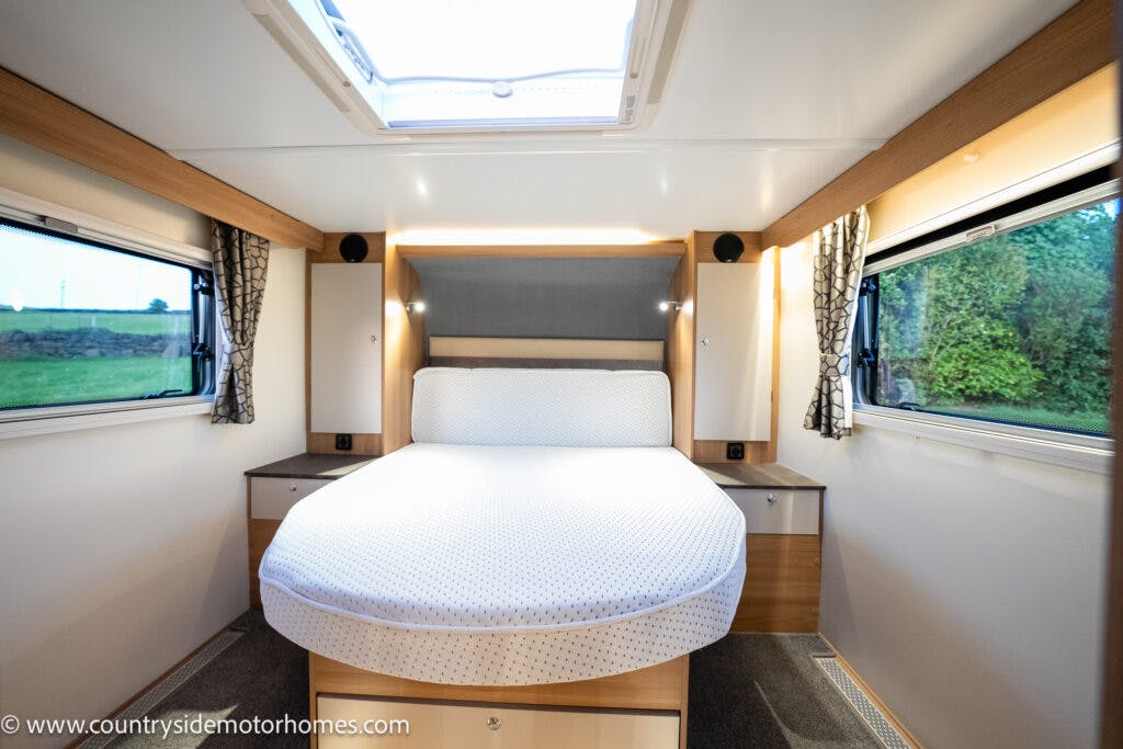 Interior of the 2021 Bailey Autograph 79-4i motorhome bedroom featuring a neatly made, round-edged bed centered. The room has two large windows with curtains on either side of the bed, nightstands, and overhead lights. A skylight is visible on the ceiling. The walls are light-colored wood.