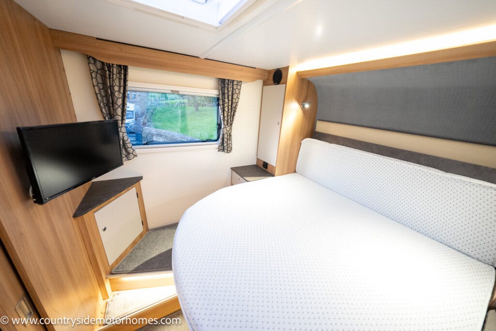 A small bedroom in the 2021 Bailey Autograph 79-4i features a fitted bed within its compact living space, complete with a wall-mounted TV, small window with curtains, and wooden decor. The room includes cabinets and drawer storage beneath the window, while the white bedding boasts a subtle pattern.