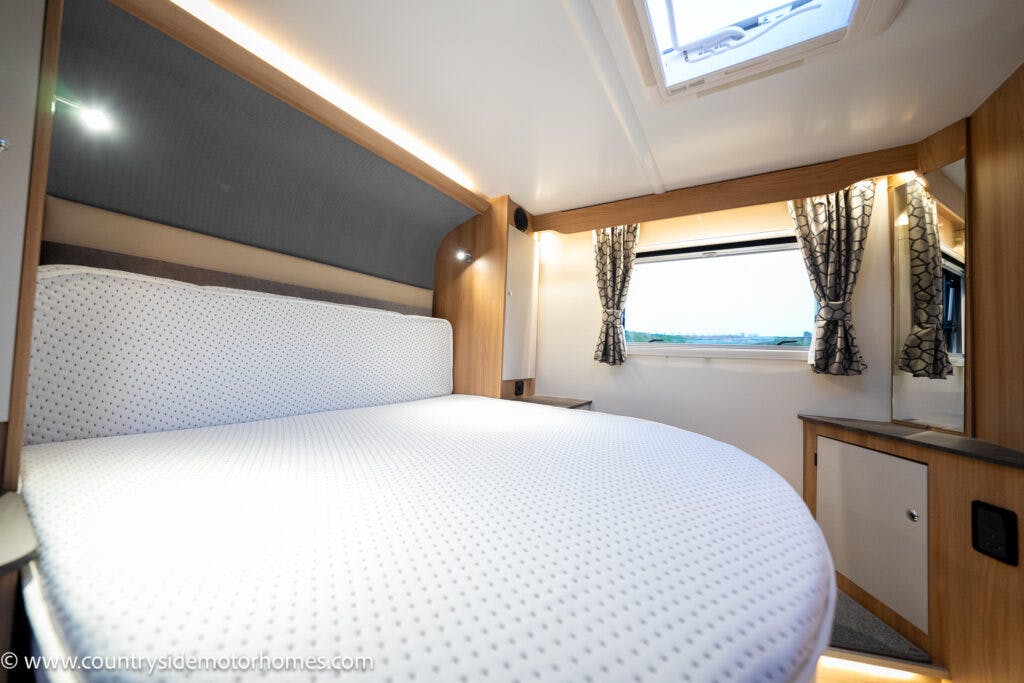 Interior of the 2021 Bailey Autograph 79-4i motorhome bedroom featuring a neatly made bed with white bedding. The room is equipped with wooden cabinetry, patterned curtains on a window, and soft lighting. A skylight is visible in the ceiling. Visit countrysidemotorhomes.com for more details.