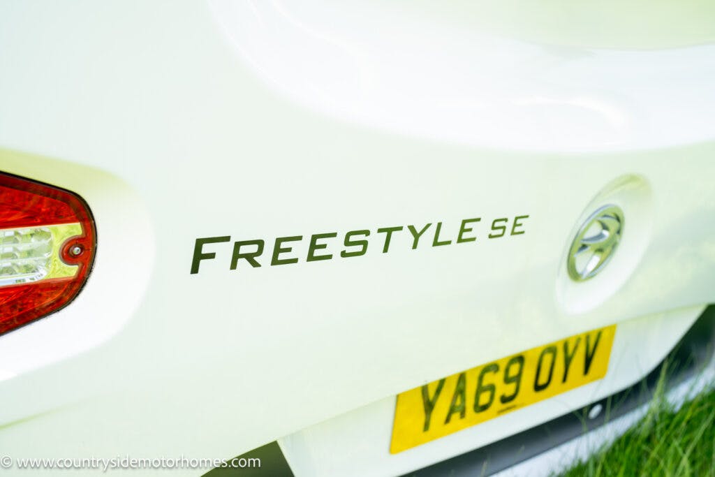 Close-up of the rear section of a white 2019 Swift Escape 694 Freestyle vehicle displaying the model name "FREESTYLE SE" in black letters. The UK license plate with yellow background and registration number "YA69 OYV" is visible below. The rear light on the left side is also partially visible.