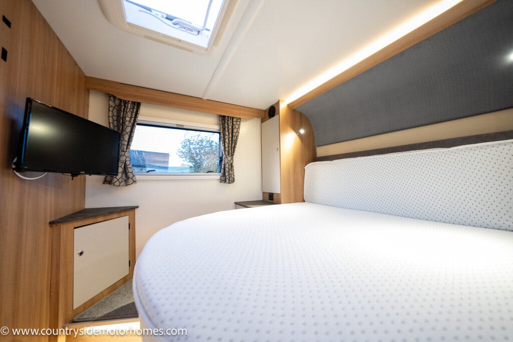 This image shows the interior of a 2021 Bailey Autograph 79-4i RV bedroom. The room features a bed with a white mattress, a wall-mounted TV opposite the bed, a window with curtains, wooden cabinetry, overhead lighting, and a skylight above the bed.
