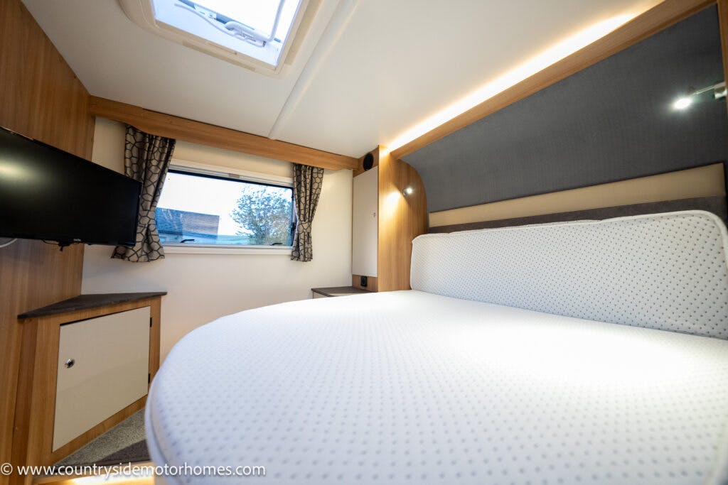 Interior view of a 2021 Bailey Autograph 79-4i motorhome bedroom featuring a neatly made bed with a rounded mattress. The room includes a wall-mounted TV, a window with curtains, and wooden cabinetry. An overhead skylight provides natural light. The website "countrysidemotorhomes.com" is visible.
