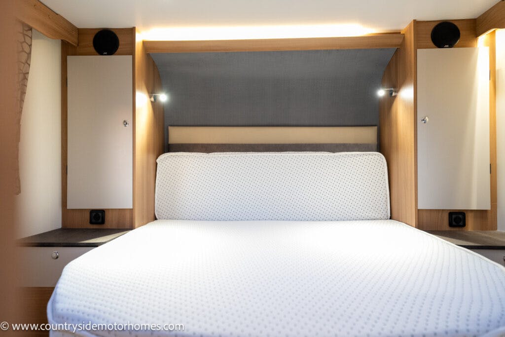 A small bedroom space inside the 2021 Bailey Autograph 79-4i features a neatly made bed with a white textured bedspread. The room has light wood cabinets and shelves on either side of the bed, integrated lighting, and dark circular elements above the cabinets.