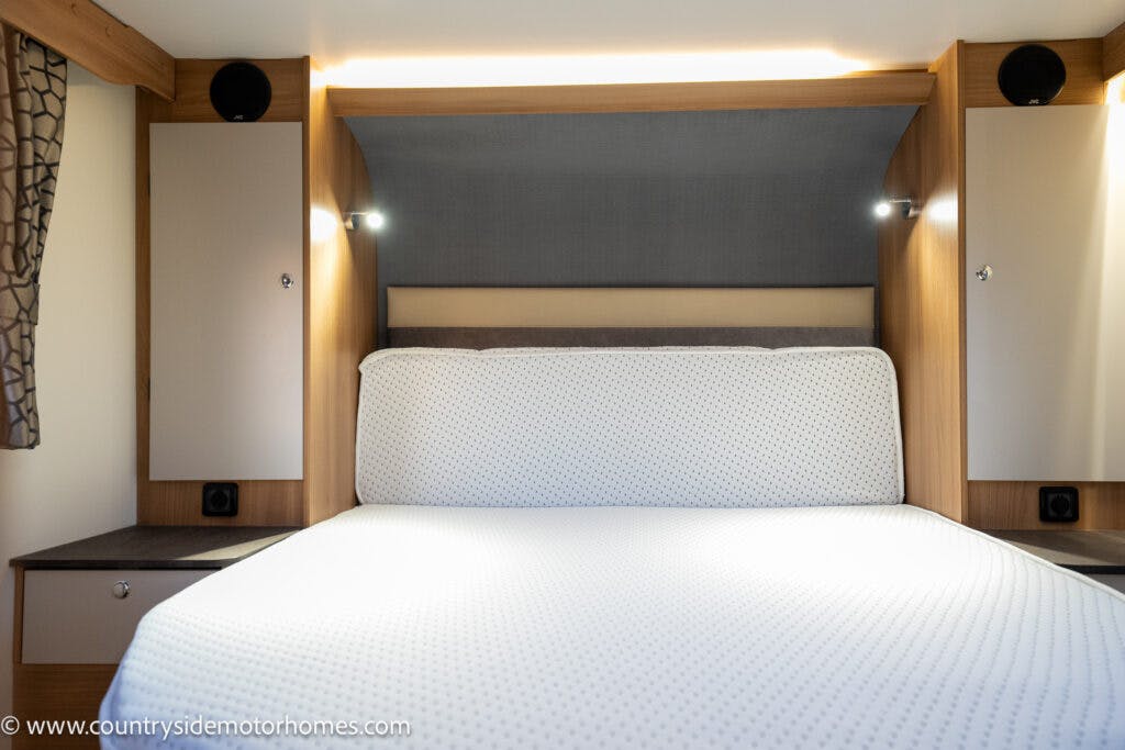 In the 2021 Bailey Autograph 79-4i, a neatly made double bed with a white patterned bedspread is centered between two wooden cabinets with overhead lights in the modern motorhome bedroom. The headboard area features a grey upholstered panel, and the light-colored walls have built-in speakers.