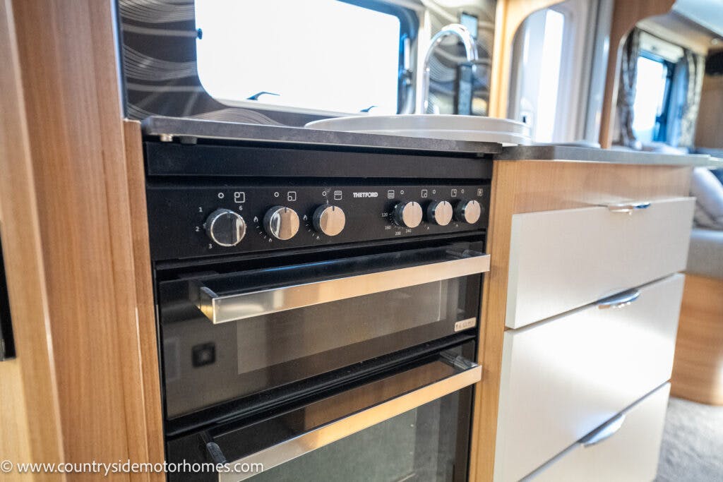 A compact kitchen inside the 2021 Bailey Autograph 79-4i motorhome featuring a built-in oven with a glass door, multiple control knobs, and a metallic handle. The oven is integrated into a wooden cabinet unit with drawers, and a sink is visible on the countertop above.