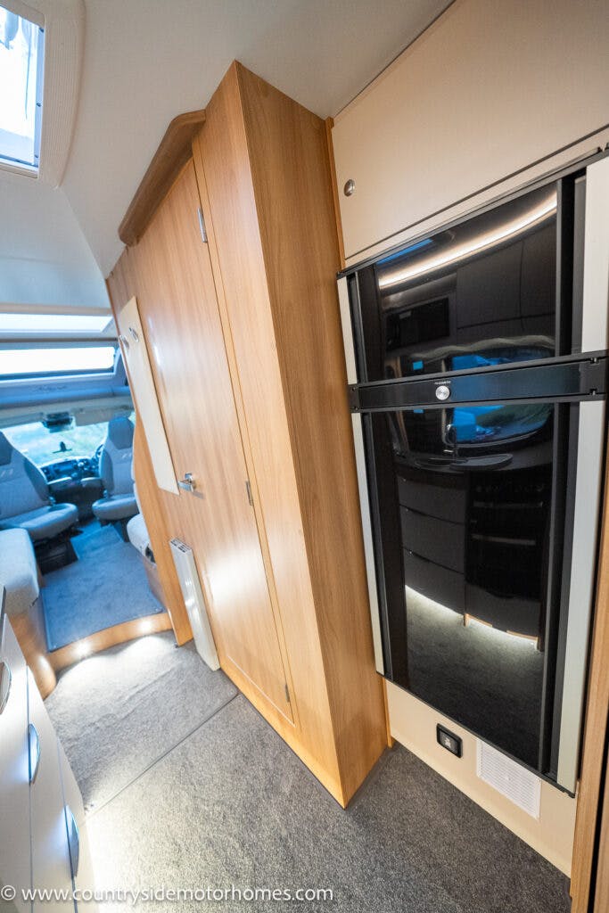 Interior of the 2021 Bailey Autograph 79-4i featuring a wooden wardrobe on the left, a black refrigerator on the right, and a glimpse of the front driving and passenger seats in the background. The motorhome has carpeted flooring and ambient lighting.