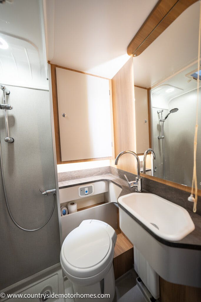 A compact bathroom inside the 2021 Bailey Autograph 79-4i motorhome. It features a white sink with a faucet, a mirror above the sink, and a toilet with storage below. The shower area has a handheld showerhead. The room is well-lit with a mix of natural and artificial light.