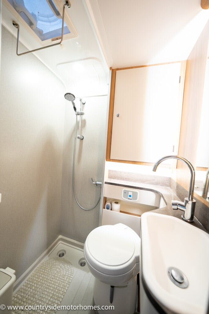 This image shows a small, compact bathroom in the 2021 Bailey Autograph 79-4i. It includes a shower with a handheld showerhead, a toilet, a sink with a faucet, and a small countertop. There is also a cabinet above the sink and a toilet paper holder near the toilet.