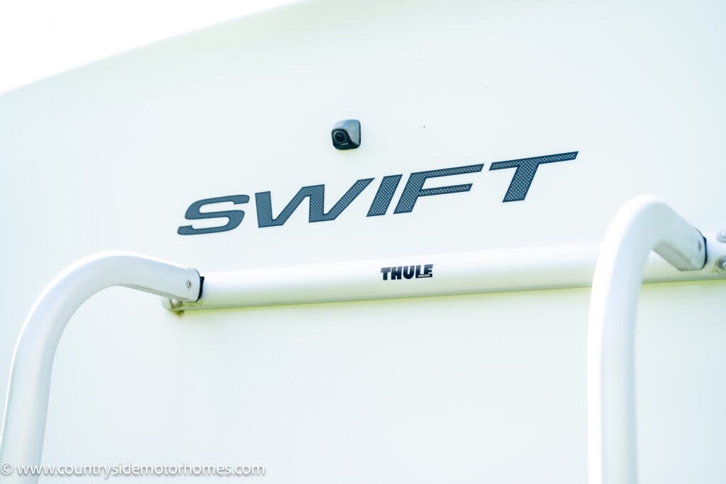 A close-up view of the back of a 2019 Swift Escape 694 Freestyle motorhome. The word “SWIFT” is prominently displayed in dark letters. Below it, there is a white Thule ladder attached to the vehicle. A small camera is visible above the word “SWIFT.”