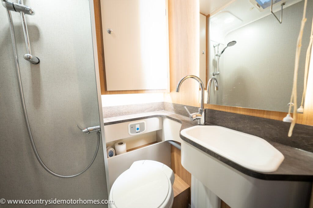 The 2021 Bailey Autograph 79-4i features a compact motorhome bathroom with a shower equipped with a flexible hose, a modern sink with a faucet, and a mirror above it. Wooden cabinets offer storage above and below the sink area, while the floor and countertop boast a dark finish for added elegance.