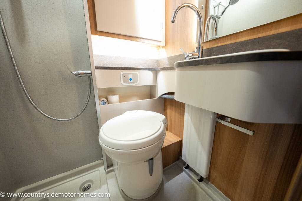 The 2021 Bailey Autograph 79-4i features a compact motorhome bathroom with a toilet, sink, and shower area. The sink has a modern faucet above a cabinet. The shower hose is mounted on the wall beside the toilet. Light wood paneling and neutral colors dominate the space, creating a serene atmosphere.