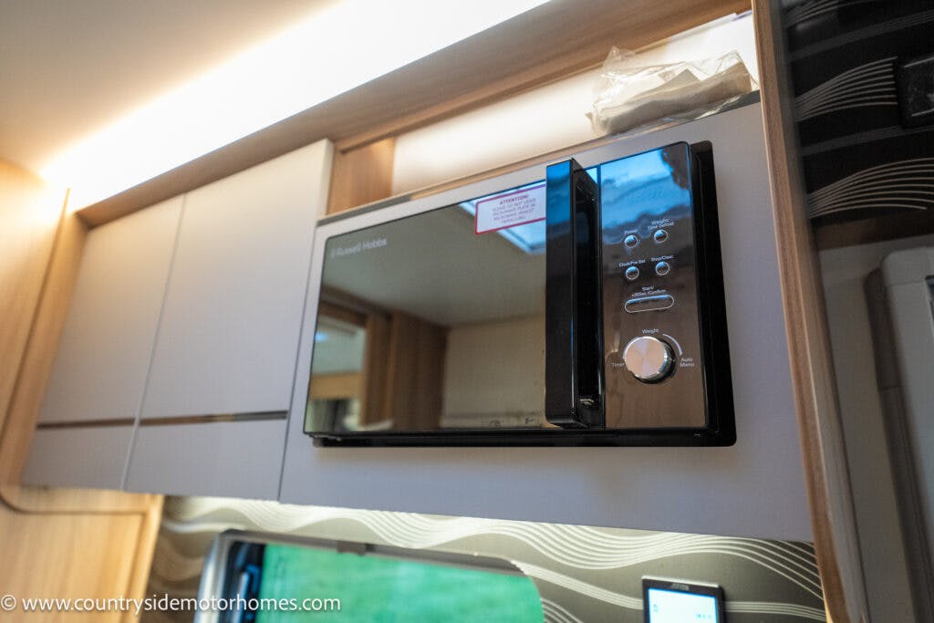 A sleek microwave oven with a black and silver front panel, seamlessly installed in a wooden cabinet above a patterned backsplash. The control panel features intuitive buttons and a dial, echoing the modern elegance found in the 2021 Bailey Autograph 79-4i.