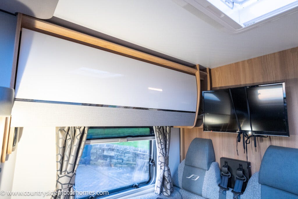 Interior view of the 2021 Bailey Autograph 79-4i motorhome featuring a light-colored overhead storage cabinet, a small window with curtains, a wall-mounted TV, and two upholstered seats with seatbelts and molded plastic backrests. Website URL is visible in the bottom left corner.