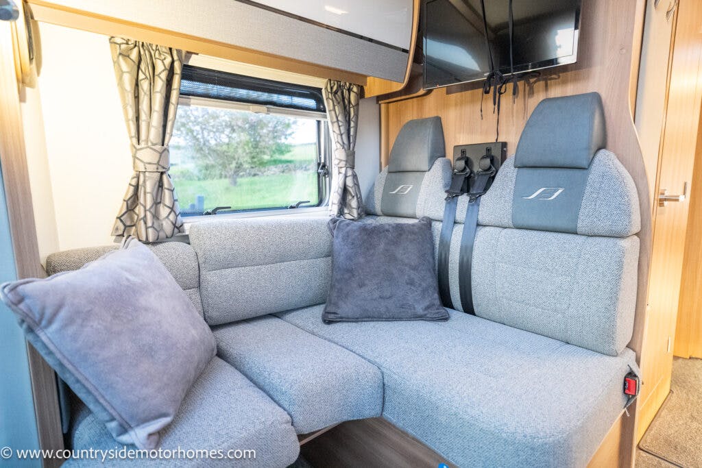 The image shows the interior of a 2021 Bailey Autograph 79-4i motorhome with gray upholstered seating arranged in an L-shape. Two seatbelt-equipped seats are positioned next to the seating area. A window with patterned curtains is visible, along with a mounted TV above the seats.