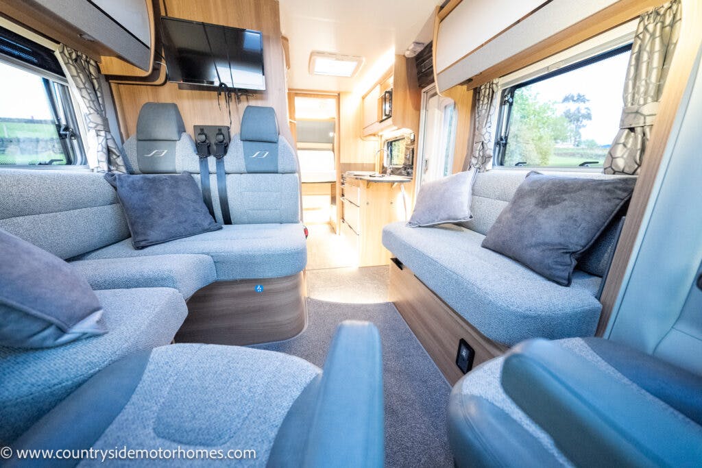 Interior of a Bailey Autograph 79-4I motorhome showcasing a seating area with two couches, grey upholstery, and multiple pillows. The space includes a small kitchen area with a sink and cupboards, overhead storage, large windows with curtains, and a ceiling vent.