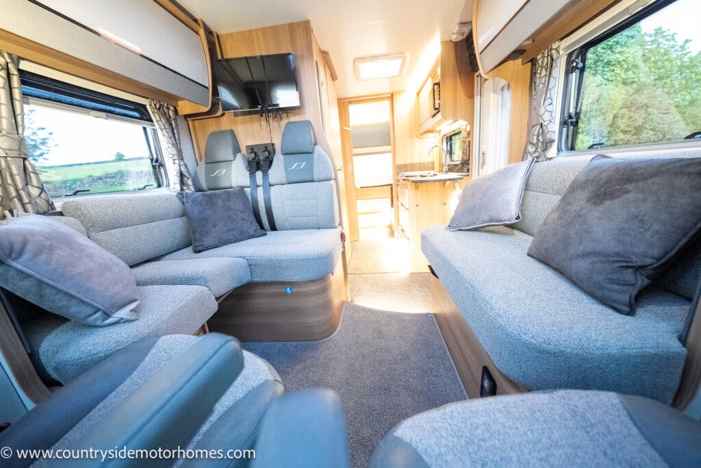 Interior of the 2021 Bailey Autograph 79-4i motorhome featuring grey cushioned seating along the sides with a dining area in the back. There are windows with curtains on either side, a kitchen area with an oven and sink, and overhead storage compartments. The floor is carpeted.