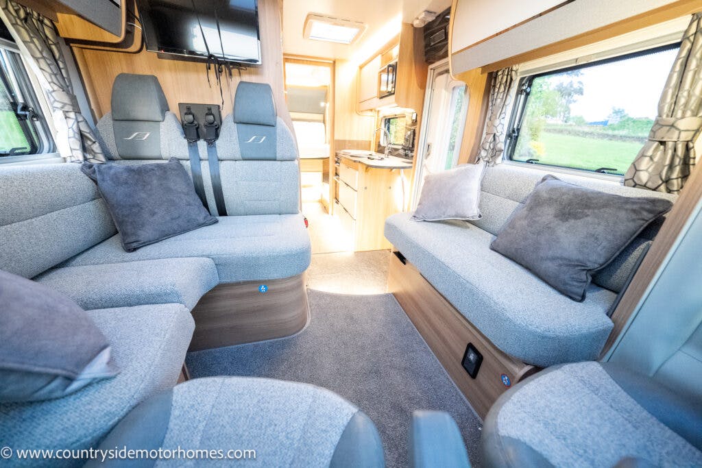The interior of the 2021 Bailey Autograph 79-4i motorhome features a lounge area with gray upholstered seating, several throw pillows, and overhead storage. A kitchenette is visible in the background with cabinets, a small sink, and a stovetop. Large windows provide natural light.
