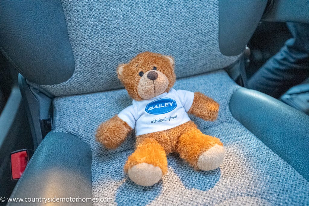 A small brown teddy bear wearing a white shirt with a blue logo and the name "Bailey" is seated on a gray fabric car seat in the 2021 Bailey Autograph 79-4i. The shirt also has the hashtag "#thebaileybear" printed on it. The teddy bear is positioned in the center of the seat.