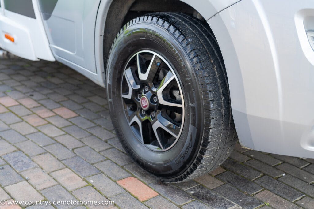 Close-up view of a motorhome's black and silver alloy wheel with a Nexen Roadian tire on a grey brick-paved surface. The 2019 Swift Escape 694 Freestyle's exterior is partially visible, showing its sleek silver body, with the source website URL displayed at the bottom left corner.
