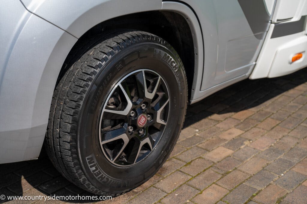 Close-up image of the front wheel of a silver 2019 Swift Escape 694 Freestyle motorhome. The wheel features a black, five-spoke alloy rim with a visible Fiat logo at the center and a Michelin tire. The motorhome is parked on a brick-paved surface in the sunlight.