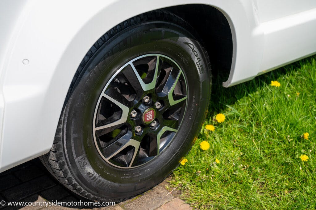 Close-up of a white 2019 Swift Escape 694 Freestyle's front wheel and tire on a driveway with grass and yellow dandelions nearby. The wheel has a black and silver design with a central logo. The image includes the URL www.countrysidemotorhomes.com.