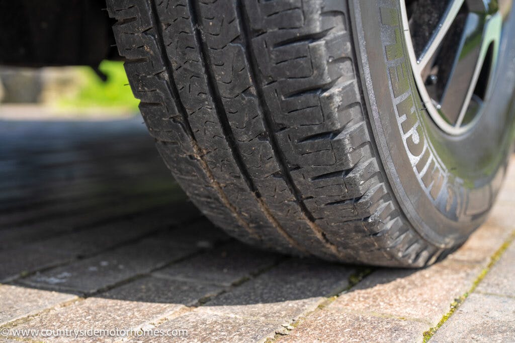 Close-up image of an automobile tire on a paved surface. The tire shows visible wear and features a distinct tread pattern. Part of the 2019 Swift Escape 694 Freestyle's wheel rim is also visible. The background includes a grassy area next to the pavement.