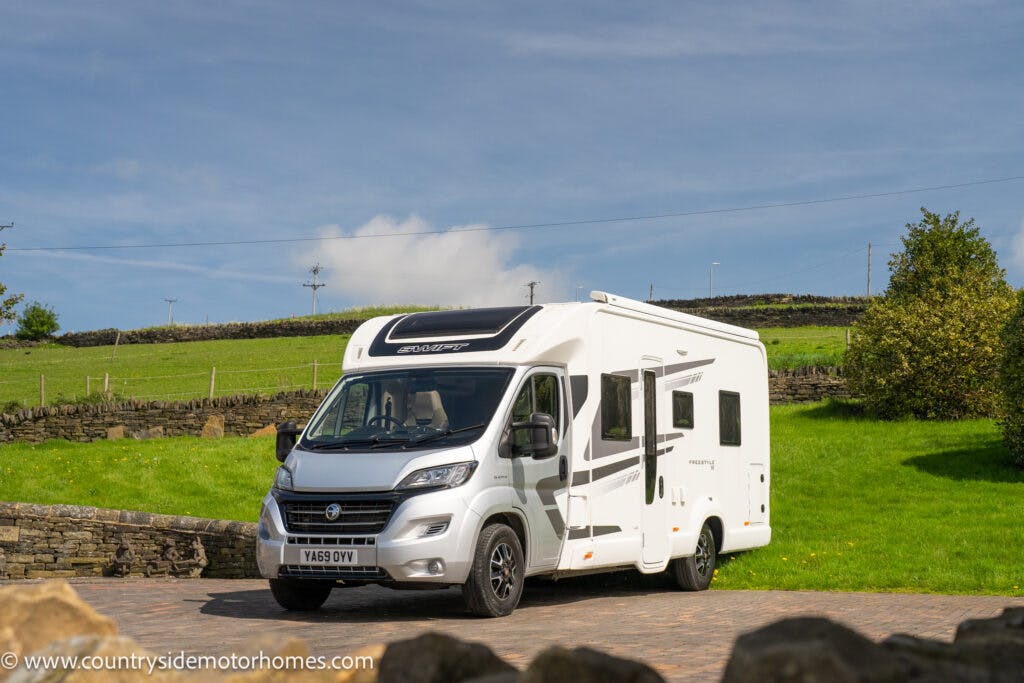 A pristine 2019 Swift Escape 694 Freestyle motorhome is parked on a paved area surrounded by green grass and stone fences. It is a sunny day with a partly cloudy sky. The vehicle features several windows, and there is a website URL on the lower left corner of the image.