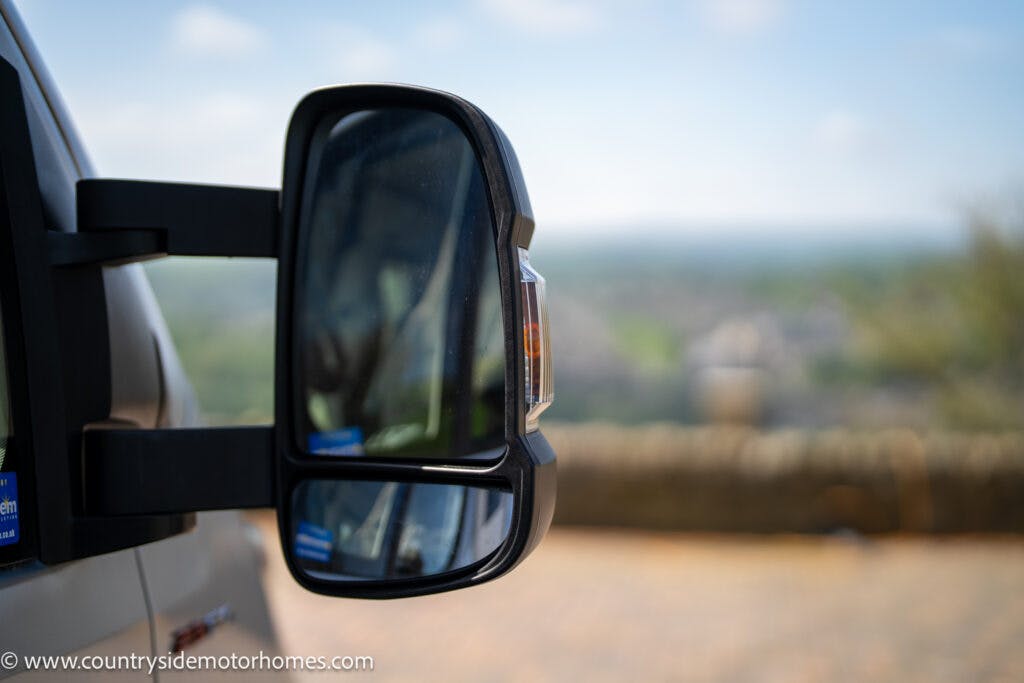 Close-up of a side mirror on a 2019 Swift Escape 694 Freestyle with a blurred outdoor background featuring greenery and a brick wall. The website URL "www.countrysidemotorhomes.com" is visible in the bottom left corner.
