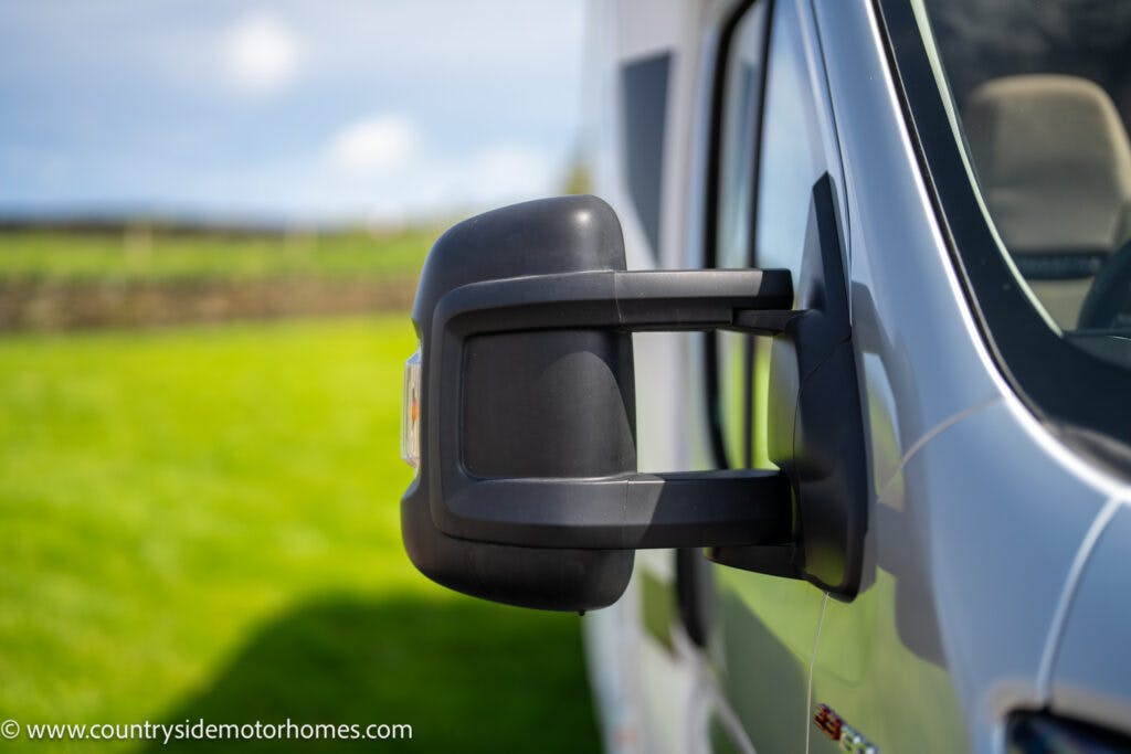 Close-up of a side mirror on a 2019 Swift Escape 694 Freestyle motorhome, with a grassy area and fields visible in the background. The image showcases the details of the mirror, with the vehicle's grey exterior partially visible. The website "www.countrysidemotorhomes.com" is in the corner.