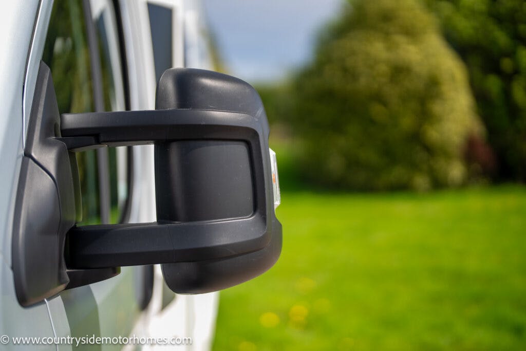 Close-up of the side mirror of a white 2019 Swift Escape 694 Freestyle, with a blurred green lawn and trees in the background. The mirror is in focus, showing its detailed structure. The vehicle is parked in a scenic, nature-filled area.