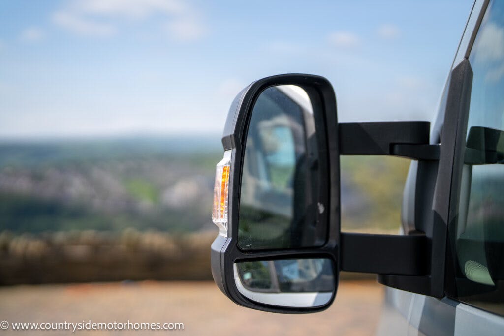 A close-up view of a side mirror on a 2019 Swift Escape 694 Freestyle motorhome with a blurred outdoor landscape in the background. The mirror reflects part of the road and scenery behind the vehicle. The motorhome is parked, and the mirror's turn signal light is visible.